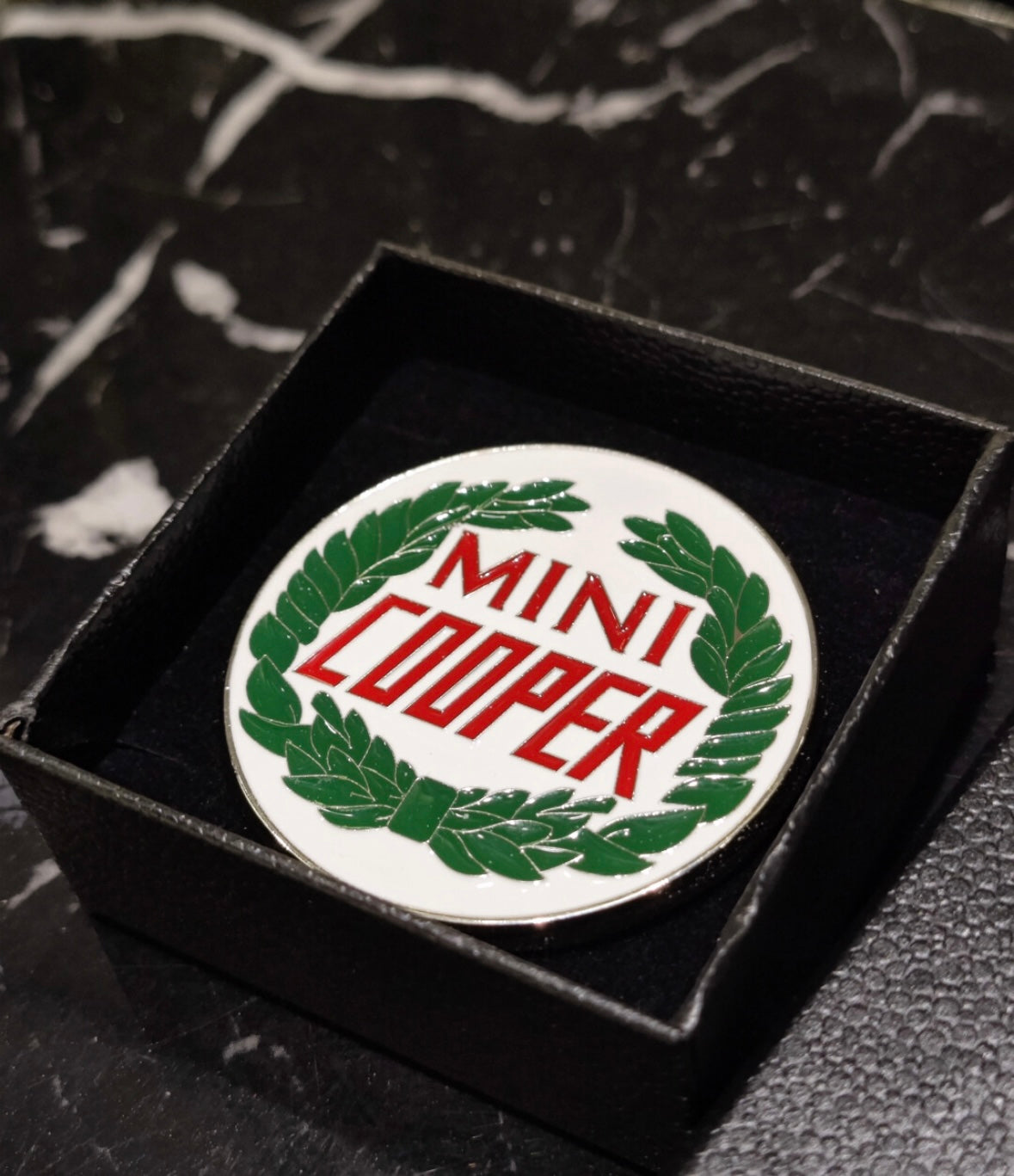 Close-up of the MINI Cooper laurel enamel badge, highlighting the detailed laurel wreath design and metal finish.